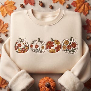 Op de afbeelding: Een crèmekleurige sweater met ronde hals en een geborduurd ontwerp van vier pompoenen op de borst. Elke pompoen is versierd met diverse bloemmotieven in tinten oranje, geel, bruin en groen, uitgevoerd met gedetailleerd stikwerk.
