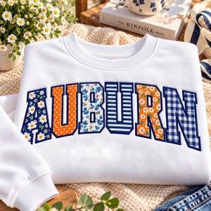 以下が含まれることがあります： 大きなブロック体の文字で「AUBURN」と書かれた白いクルーネックのスウェットシャツ。各文字には、ネイビー地に白いデイジー、オレンジのドット、青い花柄、ネイビーのストライプ、オレンジのデイジー、青いギンガムチェックなど、それぞれ異なるユニークなパターンが施されています。