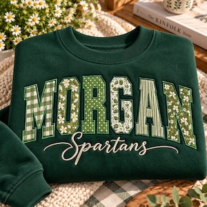 Peut inclure: Un sweat-shirt à col rond vert foncé portant le nom MORGAN en grandes lettres majuscules, chacune avec un motif unique incluant vichy, marguerites, pois et rayures. Sous le nom, le mot Spartans est brodé en écriture cursive blanche.