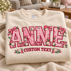 Könnte beinhalten: Ein cremefarbenes Sweatshirt mit Rundhalsausschnitt und dem Namen ANNE in großen, rosa Buchstaben mit Vichy-Muster. Die Buchstaben sind mit gestickten rosa Schleifen sowie kleinen weißen und rosa Blumen verziert. Darunter steht CUSTOM TEXT.
