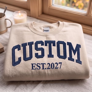 May include: A beige crewneck sweatshirt featuring dark blue embroidered lettering that reads CUSTOM EST. 2027 in a collegiate font style. The garment is folded neatly, showcasing the bold text centred on the front chest area of the fabric.