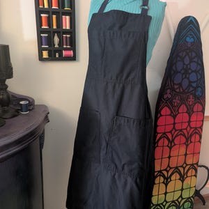 Può includere: Un grembiule blu navy scuro con due ampie tasche frontali e un cinturino per il collo. Il grembiule è esposto su un manichino. Sono visibili anche una mensola decorativa a parete con rocchetti di filo e un asse da stiro arcobaleno.