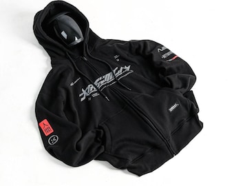 May include: A black zip-up hoodie featuring a hood with drawstrings, a front kangaroo pocket, and white graphic text on the chest. The sleeve displays a red square with Japanese characters and a circular icon, plus additional text on the upper arm.