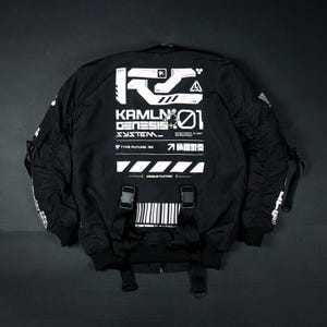 Puede incluir: Una chaqueta bomber negra con estampados gráficos blancos en la espalda, incluyendo el texto KRMLN GENESIS 01, SYSTEM y DYSTOPIA ALERT SCENARIO. La chaqueta incluye dos hebillas de plástico negro con correas colgantes y un código de barras.