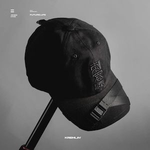 May include: A black baseball cap featuring a structured crown and a curved brim. The front panel displays black embroidered Japanese characters and the text KAS MEN. A black nylon strap with a plastic buckle crosses the brim diagonally.