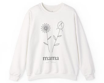 May include: A cream-colored long-sleeve sweatshirt featuring a black line-art graphic of two flowers. Below the flowers, the word "mama" is printed in a serif font, with the names "AVA" and "NOAH" positioned underneath in smaller text.