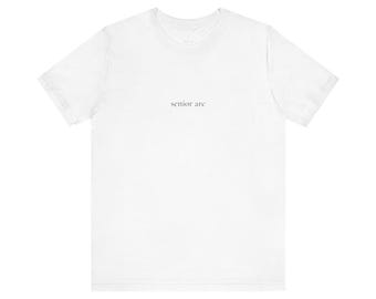 May include: A plain white short-sleeved t-shirt featuring the text "senior arc" printed in small, centered, dark grey lowercase letters on the chest area. The garment has a standard crew neckline and a simple, clean design throughout the fabric.