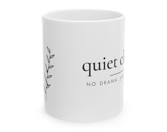 May include: A white ceramic mug featuring a black line drawing of a leafy branch on the left side. The text on the mug reads quiet c, with the phrase NO DRAMA. JUST printed in smaller capital letters underneath the main text in a black font.