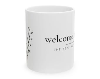 May include: A white ceramic mug featuring a black line drawing of a leafy branch on the left side. The text "welcome home" is printed in a black serif font, with the text "THE KEYS ARE" positioned directly underneath in a smaller sans-serif font.