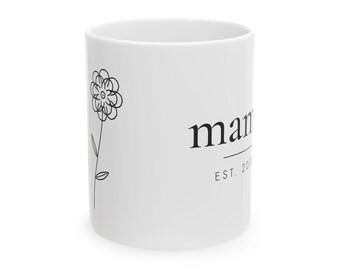 May include: A white ceramic mug featuring a black line drawing of a flower on the left side. The text "mama" is printed in a black serif font on the right, with the text "EST. 201" positioned directly below it, separated by a thin horizontal line.