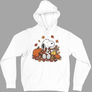 May include: A white hooded sweatshirt featuring a graphic print of Snoopy and Woodstock sitting on a plaid blanket with pumpkins and falling autumn leaves. The characters are wrapped in a matching plaid scarf, creating a seasonal autumn theme.