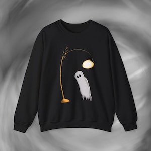 Può includere: Una felpa nera a girocollo con una stampa grafica raffigurante un piccolo fantasma grigio chiaro in piedi sotto una lampada da terra alta, sottile e color giallo oro, con un paralume arrotondato e luminoso. Il disegno è centrato sul davanti.