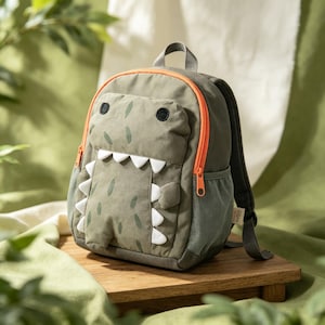 May include: An olive green backpack designed to resemble a dinosaur, featuring a front pocket with black embroidered eyes, white fabric teeth along the zip, and small green spots. The bag includes bright orange zip trim and mesh side pockets.