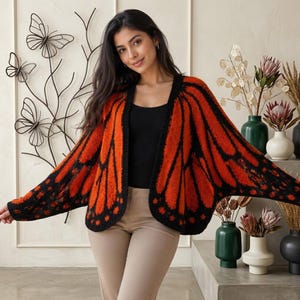 May include: A knitted open-front cardigan featuring a vibrant orange and black monarch butterfly wing pattern. The garment has long, wide sleeves, a solid black trim along the front opening, and a soft, textured knit construction.