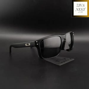 May include: Matte black sunglasses featuring dark tinted lenses and a rectangular frame shape. The temples display a silver-toned rectangular logo emblem near the hinges, with two small silver-toned rivets on the front frame corners.