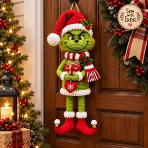 May include: A green crocheted character decoration wearing a red and white Santa hat, a matching striped scarf, and red curled-toe boots. The figure holds a small red gift box and a red heart ornament with a white snowflake design on a string.