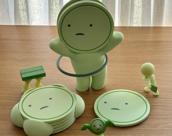 May include: A collection of light green Smiski figures and accessories, including a large figure with a hula hoop, a stack of circular coasters with faces, a small green bench with the text SMISKI, and two smaller character figurines.