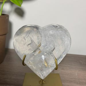Pode incluir: Um cristal de quartzo transparente em forma de coração com fraturas e inclusões internas visíveis, montado num suporte de metal dourado com base quadrada. O cristal é transparente e incolor, exibindo texturas cristalinas naturais.