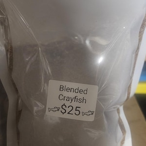 May include: A clear, resealable plastic pouch containing a finely ground, dark-colored powder. A white rectangular label is affixed to the front, featuring the text Blended Crayfish with a small fish graphic on either side of the price.
