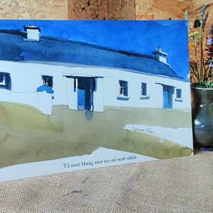 May include: Watercolour painting of a white cottage with a blue roof and doors under a bright blue sky. The artwork includes the text "Tá nead bheag níos teo ná nead mhór."