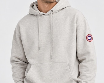 May include: A light gray heathered long-sleeve hooded sweatshirt featuring a front kangaroo pocket, a drawstring hood with metal aglets, and a circular red, white, and blue logo patch on the upper left sleeve, paired with black trousers.