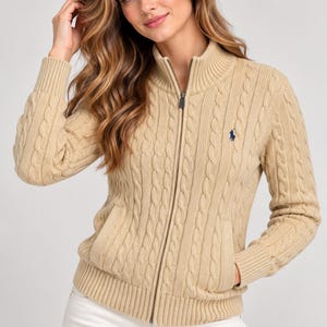 May include: A beige cable-knit zip-up sweater featuring a mock neck collar, long sleeves with ribbed cuffs, and two front pockets. The garment includes a small navy blue embroidered logo on the left chest and a full-length metal zipper closure.