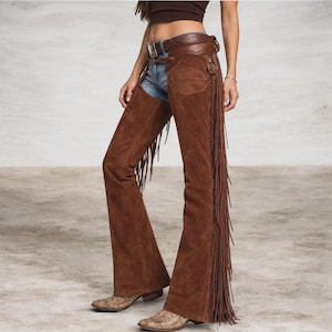 May include: Brown suede leather chaps featuring long fringe detailing along the outer legs, a metal buckle closure at the waist, and decorative metal studs. The chaps are worn over blue denim jeans and paired with light brown patterned leather boots.