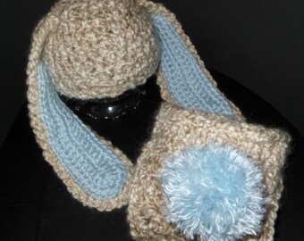 May include: A crocheted hat and scarf set in beige and light blue. The hat has a pom-pom on top and the scarf has long, floppy ears.