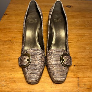 Pode incluir: Um par de sapatos de salto Stuart Weitzman com um padrão de pele de cobra texturizado em cinzento e castanho. Cada sapato tem uma biqueira quadrada e uma fivela decorativa em tom de latão antigo na frente. O forro interior é dourado.