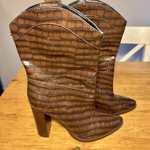 Könnte beinhalten: Ein Paar wadenhohe braune Stiefel mit einer strukturierten Krokodilprägung. Die Stiefel haben eine spitz zulaufende Zehenpartie, einen stabilen Blockabsatz und ein dreieckiges Ziernahtdetail am Schaft sowie eine glatte braune Sohle.