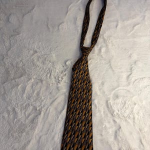 Könnte beinhalten: Eine Krawatte mit einem sich wiederholenden geometrischen Muster aus winkligen Formen in Senfgelb, Anthrazit und Schwarz. Die Krawatte ist oben geknotet, wobei das lange Ende nach unten verläuft und ein durchgehendes dreieckiges Motiv zeigt.
