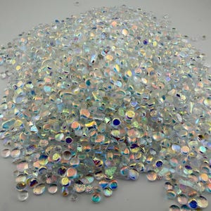 May include: A large collection of small, clear glass pebbles with an iridescent finish. Each pebble features a smooth, rounded shape and reflects light with multi-colored highlights including blue, pink, and gold tones across the surface.