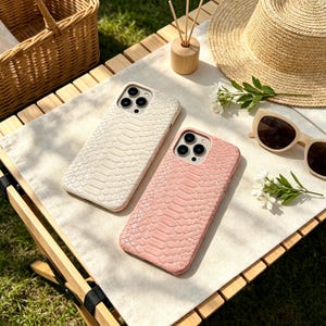 Op de afbeelding: Twee smartphonehoesjes met een gestructureerd slangenleerpatroon. Het ene hoesje is gebroken wit en het andere is lichtroze. Beide hoesjes hebben uitsparingen voor een drievoudig camerasysteem en liggen op een plat oppervlak.