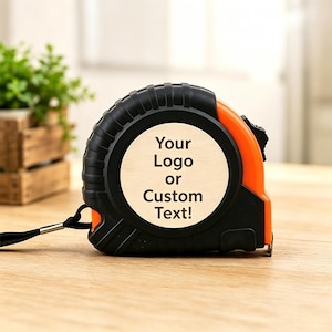Könnte beinhalten: Ein schwarzes und orangefarbenes Rollmaßband mit einer runden Holzeinlage in der Mitte. Auf der Holzfläche ist der schwarze Text "Your Logo or Custom Text!" aufgedruckt. Das Gerät verfügt über eine seitlich befestigte schwarze Handschlaufe.