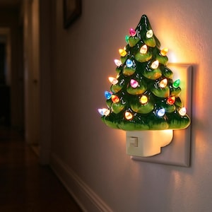 Puede incluir: Una luz nocturna de árbol de Navidad de cerámica verde enchufada a una toma de corriente. El árbol tiene un acabado brillante y está decorado con pequeñas bombillas brillantes de colores rojo, azul, amarillo y blanco.