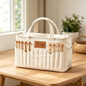 Op de afbeelding: Een crèmekleurige rechthoekige opbergtas met twee handvatten en een ritssluiting. De voorzijde heeft een bruine patch in lederlook met de tekst "Knit HAPPY" en verticale vakken waarin diverse houten breinaalden zijn opgeborgen.