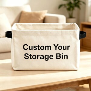 May include: A rectangular off-white canvas storage bin featuring black text that reads Custom Your Storage Bin on the front. The bin has two black fabric handles attached to the sides and a structured, open-top design for organizing household items.