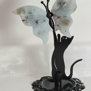 May include: A black metal cat figurine standing on a circular base, reaching upward with one paw to hold a translucent, pale blue butterfly ornament featuring subtle dark speckles and reddish inclusions within its wings.