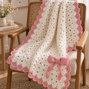 Könnte beinhalten: Eine cremefarbene Häkeldecke mit einem zarten Lochmuster und einem gewellten rosa Rand. Die Decke ist mit einer auffälligen, strukturierten rosa Häkelschleife verziert, die am unteren Teil befestigt ist und ein dekoratives Element darstellt.