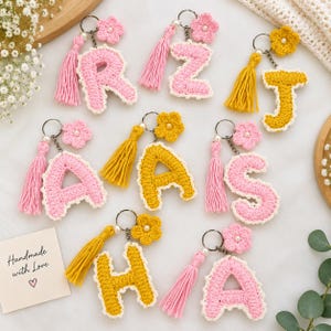 Puede incluir: Una colección de llaveros con letras de ganchillo en hilo rosa y amarillo. Cada letra tiene un borde blanco, una borla de hilo a juego y una pequeña flor de ganchillo con una cuenta central. Se incluye una tarjeta con el texto Handmade with Love.