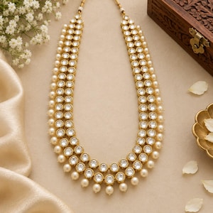May include: A double-strand necklace featuring round, clear, faceted stones set in gold-toned metal. The inner and outer strands are lined with small, cream-colored spherical beads, with additional beads dangling from the bottom edge of the necklace.