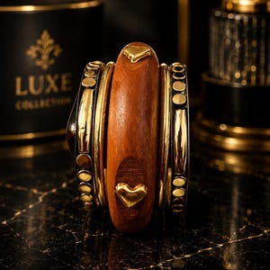 May include: A stack of bangles featuring a central thick wooden bracelet with two small gold-toned heart-shaped accents. Flanking the wooden piece are two thinner black metal bangles decorated with a series of small, circular gold-toned studs.