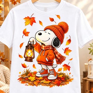 May include: A white short-sleeved t-shirt featuring a graphic of a cartoon dog wearing an orange knit hat and scarf, holding a glowing lantern while standing in a pile of colorful autumn leaves with falling foliage surrounding the character.