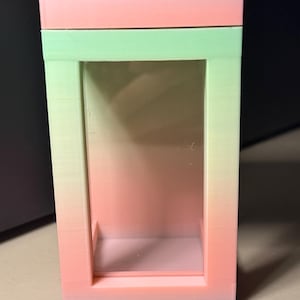 Puede incluir: Una caja de exhibición rectangular con un esquema de color degradado que transita de un tono melocotón suave en la parte superior e inferior a un verde pálido en el centro. La caja tiene un panel frontal transparente y reflectante con un marco sólido de capas horizontales visibles.