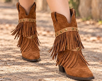 May include: A pair of brown suede ankle boots featuring two horizontal straps adorned with metallic studs and long fringe detailing. The boots have a pointed toe, a stacked block heel, and a deep V-shaped cutout at the top of the shaft.