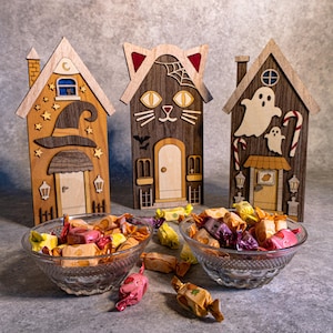 Puede incluir: Tres adornos decorativos de madera con forma de casa que presentan un sombrero de bruja, una cara de gato y fantasmas, exhibidos detrás de dos cuencos de cristal llenos de caramelos envueltos en colores como amarillo, rosa y marrón.