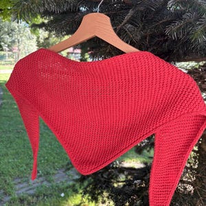 May include: A bright red triangular shawl featuring a textured knit pattern. The garment is displayed on a wooden hanger, showcasing its wide top edge and two long, pointed ends that drape downward in a symmetrical, angular shape.