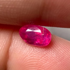 Puede incluir: Una gema de corte ovalado con un tono rosa rojizo profundo y saturado. La piedra presenta una superficie facetada que refleja la luz, mostrando su claridad interna y un acabado pulido. La gema se sostiene para mostrar su color.