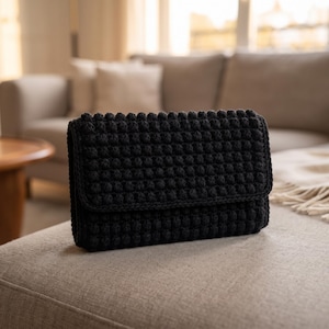 May include: A rectangular black clutch bag featuring a dense, raised bobble stitch crochet texture. The bag has a structured flap closure that covers the front panel, showcasing a uniform, repetitive pattern of small, rounded yarn knots.