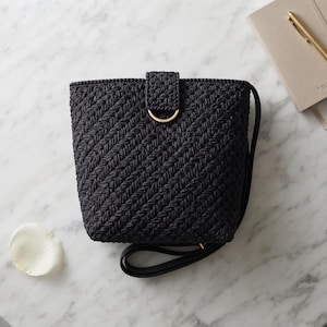 May include: A black bucket bag with a textured, diagonal crochet pattern. The bag features a central vertical strap secured by a gold-toned D-ring hardware piece at the top center. A black shoulder strap is attached to the side of the bag.
